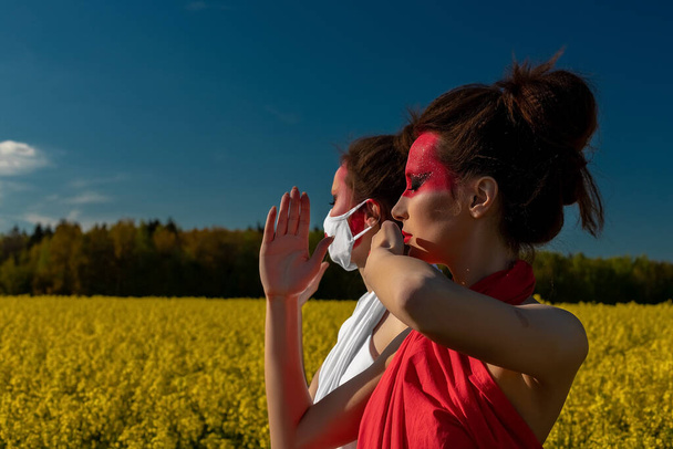Two beautiful young brunette girls with creative bright makeup in tunics on a background of a field of yellow flowers and blue sky. One girl in a mask, the other girl closes her mouth with her hand. - Фото, изображение
