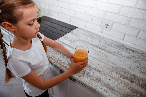 Little girl drinks fresh orange juice from a glass cup in the kitchen - 写真・画像