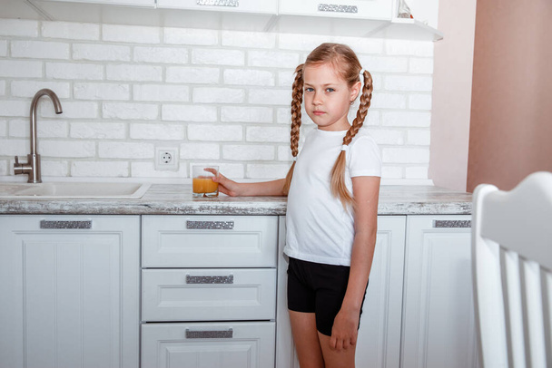 Little girl drinks fresh orange juice from a glass cup in the kitchen - Valokuva, kuva