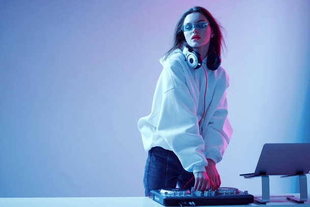 Cool young girl DJ mixes music on a mixing console and laptop, in stylish clothes, glasses on a neon background. - Fotografie, Obrázek