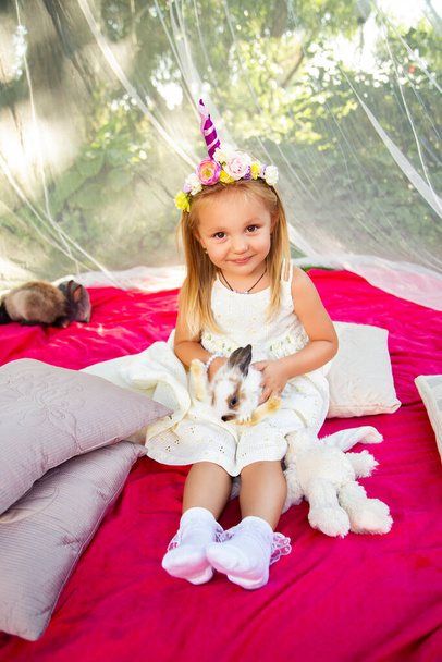 Cute little girl on a pink coverlet in nature in a dress and a unicorn hoop with rabbits - Foto, Imagem