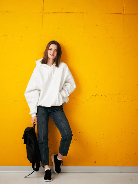 Portrait of a young pretty girl student in a white hoodie, casual clothes, with a bag, against a concrete orange wall. - Fotografie, Obrázek