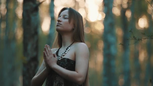 jonge vrouw bidden met gesloten ogen in de natuur tijdens de zonsondergang omgeven door bomen - Video