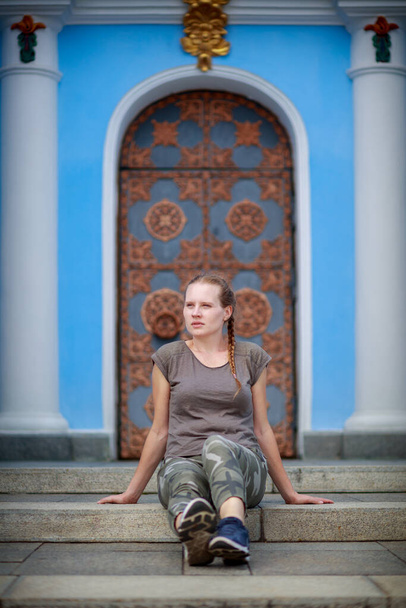 Μια όμορφη γυναίκα κάθεται στα σκαλιά. Ένας μπλε τοίχος και όμορφες πόρτες στο βάθος. Γυναίκα με σκούρο καφέ μπλουζάκι και χακί στρατιωτικό παντελόνι.. - Φωτογραφία, εικόνα