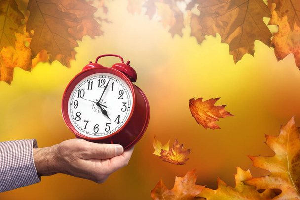 Man's hand holding alarm clock, autumn leaves background, daylight saving time concept - Photo, Image