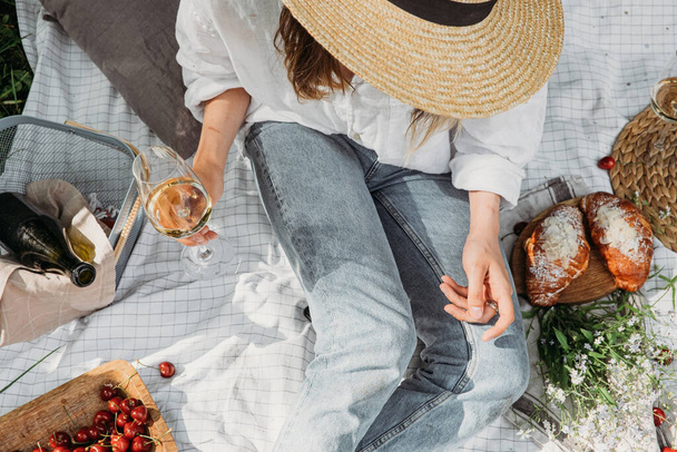 Woman in straw hat on picnic holding a glass of wine Top view. Aesthetic picnic outdoors with wine and berries and croissants.  - Valokuva, kuva