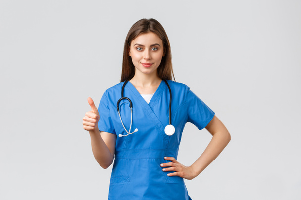 Medical workers, covid-19 and vaccination concept. Confident professional doctor, female nurse in blue scrubs and stethoscope, show thumbs-up, assure or guarantee best quality of service in clinic - Photo, Image