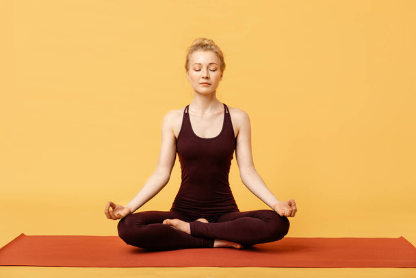 Mulher loira jovem meditando, fazendo ioga postura de lótus padmasana e asana. Menina fitness desfrutando de ioga em ambientes fechados em roupas esportivas em fundo amarelo, isolado. Prática de meditação ioga sem estresse
 - Foto, Imagem
