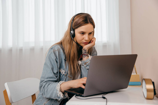 Mujer joven freelance enfocada sentada en la mesa en casa con laptop y auriculares, trabajando remotamente en línea desde casa. Adolescente concentrada estudiando a distancia sola en la sala de estar. - Foto, Imagen