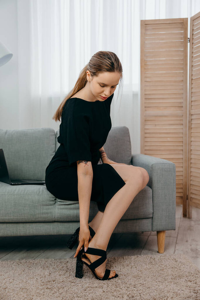 Уставшая молодая деловая женщина в черном платье сидит на диване и снимает обувь - Фото, изображение