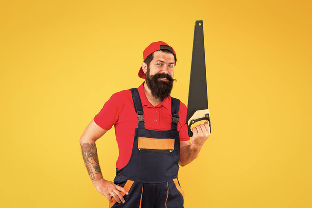 He loves his job. engineer worker career. brutal male worker with saw. man builder yellow background. professional repairman in uniform. build and construction. Skilled architect repair and fix - Fotografie, Obrázek