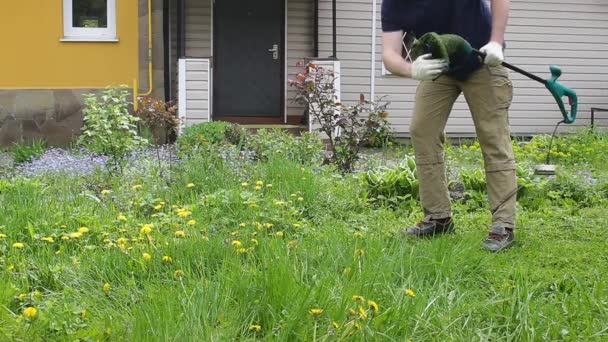 Een mannelijke tuinman reinigt een elektrische trimmer tijdens het werken in de tuin, van ruw gras. Zomerwerk van gemeentelijke diensten en particuliere werknemers. - Video