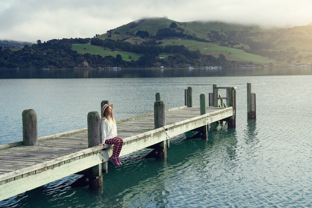 Όμορφη Ασιάτισσα που ξεκουράζεται κοιτάζει τον κόλπο της θάλασσας με τα σπίτια και τις βάρκες σε μια ηλιόλουστη μέρα στο Childrens Bay, Akaroa, Canterbury, Νέα Ζηλανδία. Χαλαρωτική νεαρή γυναίκα στο λιμάνι. - Φωτογραφία, εικόνα