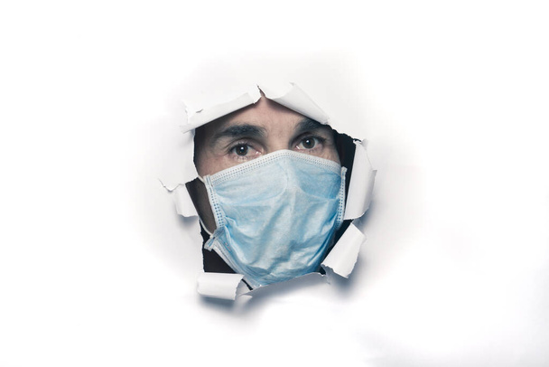 Keski-ikäinen valkoihoinen mies yllään suojaava naamio tai kirurgi murtaa valkoinen paperi arkki tai seinä katsomassa ulkopuolella vankeudessa coronavirus covid-19 - Valokuva, kuva