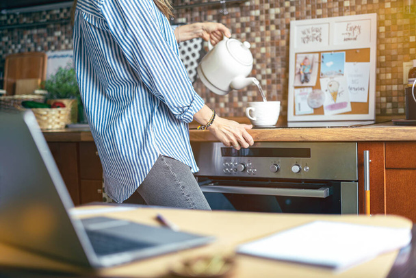 Young cropped woman preparing tea at wooden table in kitchen. Pouring water into a cup from kettle before job. Distance learning online education and work at home with computer. - 写真・画像