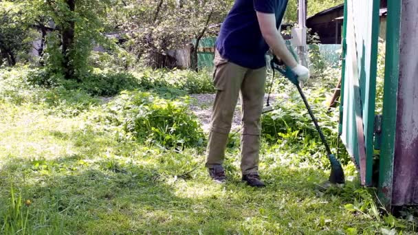 A tall man in a blue t-shirt and medical mask mows the lawn with an electric lawn mower. Warm summer weather, grass moving in the wind. Weed and dandelion control in the garden of a private house. - Footage, Video