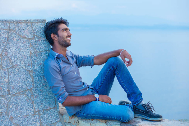 Man smiling looking up to blue sky taking deep breath celebrating freedom while sitting on a concrete wall bridge above the sea. Positive person, peace mind concept. Free happy guy enjoying sunset - Foto, Imagem