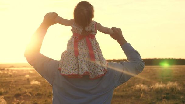 Šťastné zdravé dítě si hraje se svým otcem na hřišti v paprscích teplého západu slunce. Táta se svou milovanou dcerou na ramenou tancuje za letu a směje se. otec a dítě cestovat v parku v létě. - Fotografie, Obrázek