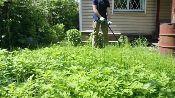 Ένας αρσενικός κηπουρός καθαρίζει ένα ηλεκτρικό χορτοκοπτικό ενώ εργάζεται στην αυλή, από ακατέργαστο γρασίδι. Θερινή εργασία δημοτικών υπηρεσιών και ιδιωτικών υπαλλήλων. - Πλάνα, βίντεο