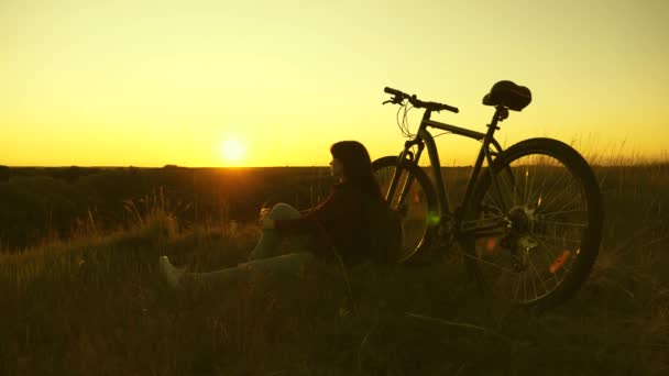 Müde Radfahrerin, die sich abends am Rand der Klippe ausruht und die Sonne bewundert. Kostenlose Mädchen reist mit dem Fahrrad bei Sonnenuntergang. Gesunde junge Wanderin sitzt auf einem Hügel neben einem Fahrrad und genießt Natur und Sonne - Filmmaterial, Video
