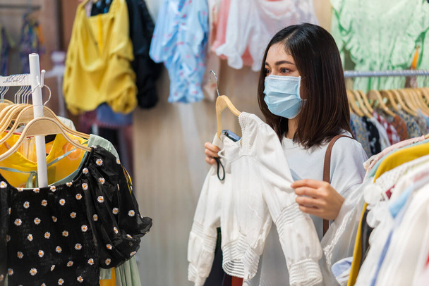mujer joven que elige ropa en el centro comercial y su uso de máscara médica para la prevención de la pandemia del coronavirus (Covid-19). nuevos conceptos normales - Foto, Imagen