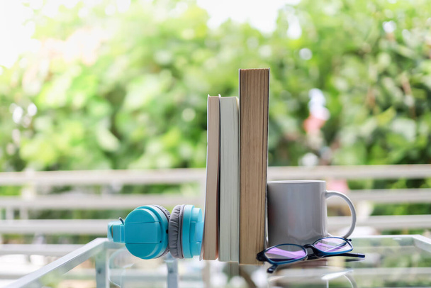 Bildungs- und Entspannungskonzept. Nahaufnahme einer weißen Tasse heißen Kaffees, hellblauem Kopfhörer, Lesebrille und Büchern auf einem Glastisch im grünen Gartenblick - Foto, Bild