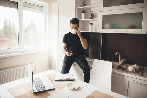 Young caucasian programmer is having a break drinking a coffee and chatting on phone in the kitchen - Photo, Image