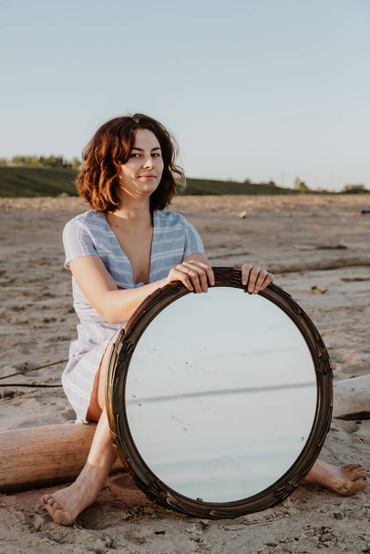 Szabadtéri hangulatos életmód fotó fiatal gyönyörű sötét hajú nő nyári ruha pózol a strandon a tükröződés a tükör.Fogalmi fotó egy tükörrel. - Fotó, kép