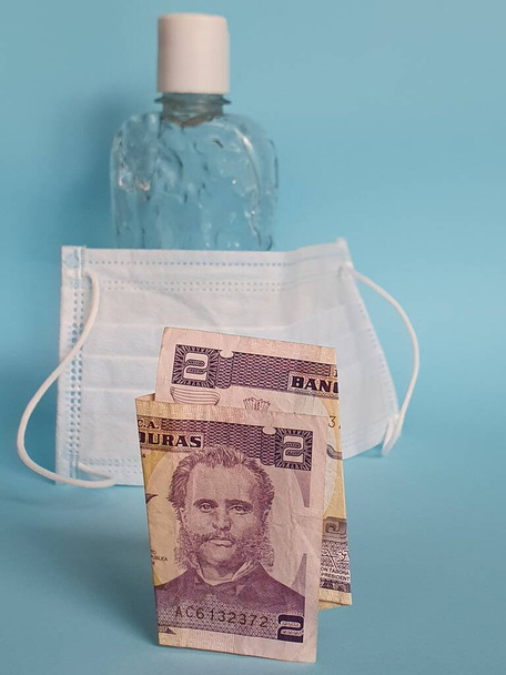 Hondurese bankbiljet van twee lempiras, gezichtsmasker, fles met gel alcohol en blauwe achtergrond - Foto, afbeelding