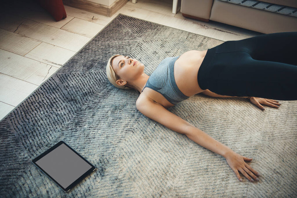 Blonde sporty woman wearing sportswear is doing fitness exercises on the floor using a tablet - Zdjęcie, obraz