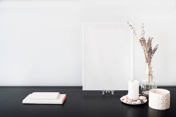 Blank white canvas for artist's mock-up, glass bottle vase with dry flowers bouquet, candles, white books on black table and white wall. Minimalism style. - Photo, image