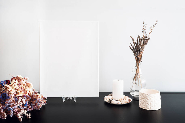 Blank white canvas for artist's mock-up, dry flowers bouquet, candles, white books on black table and white wall. Minimalism style. - Photo, Image