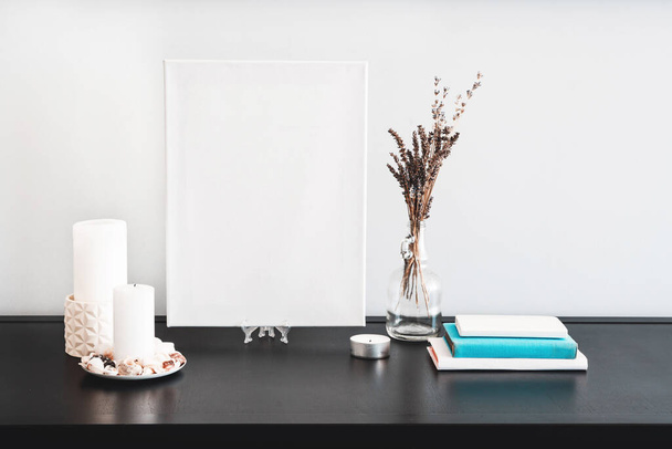 Blank white canvas for artist's mock-up, glass bottle vase with dry flowers bouquet, candles, white books on black table and white wall. Minimalism style. - Photo, Image