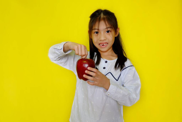 A cute young Asian girl holding up an apple, getting ready to eat it for breakfast. Bright yellow background. - Foto, Imagen