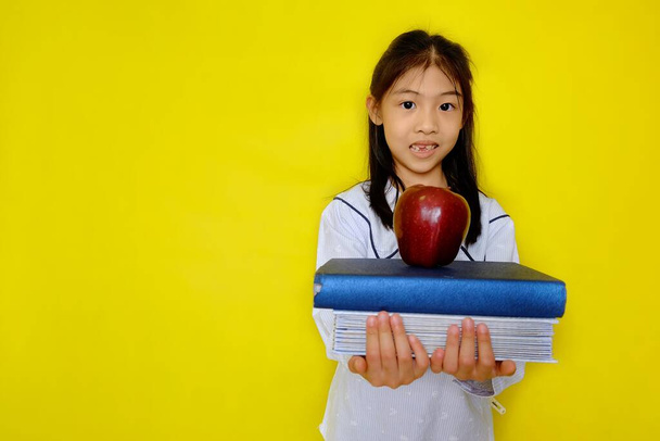 Una linda joven asiática sosteniendo una pila de libros con una manzana roja en la parte superior preparándose para volver a la escuela. Fondo amarillo brillante
. - Foto, Imagen
