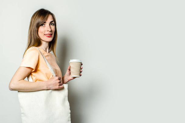 Jeune femme avec un sac à provisions et tient une tasse en papier avec du café sur un fond clair
. - Photo, image