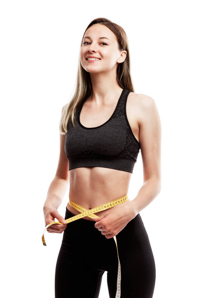 Slender laughing young girl in black sportswear measures her waist. Diet and exercise. Isolated on a white background. Vertical. - Photo, image