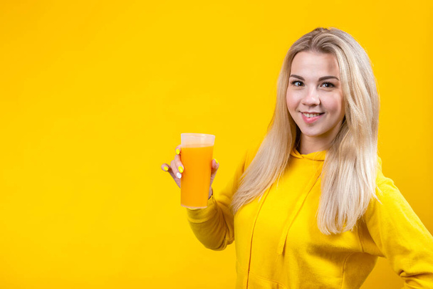 Vrolijke mooie jonge blonde vrouw in geel casual sportieve kleding met sinaasappelsap glas, lachend meisje geïsoleerd op gele achtergrond, op zoek naar camera. - Foto, afbeelding