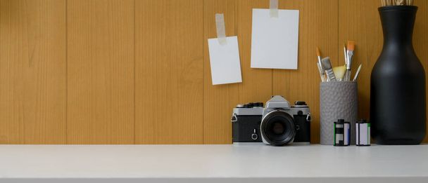 Közelkép az egyszerű munkaterület fényképezőgéppel, ecsettel, vázákkal és másolás helyet fehér asztalon jegyzettömbbel a deszka falán - Fotó, kép
