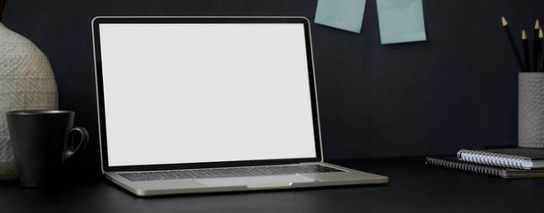 Обрезанный снимок темного современного рабочего пространства с макетом ноутбука, канцелярских принадлежностей, кружки и украшения на черном столе с блокнотом на стене
 - Фото, изображение