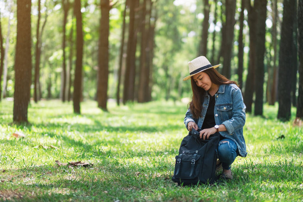 Eine asiatische Reisende mit Hut öffnet ihren Rucksack, während sie in einem schönen Kiefernwald unterwegs ist - Foto, Bild