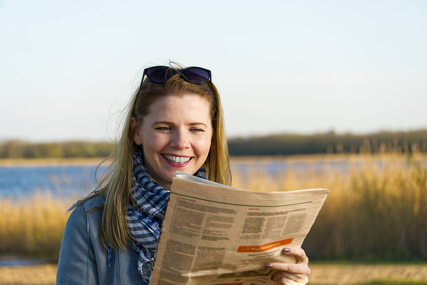 szczęśliwa kobieta czytająca gazetę nad jeziorem. dobra koncepcja wiadomości. - Zdjęcie, obraz