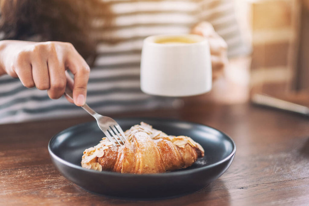 Imagen de cerca de una mujer comiendo un trozo de croissant por tenedor para el desayuno con taza de café en la mesa de madera
  - Foto, Imagen