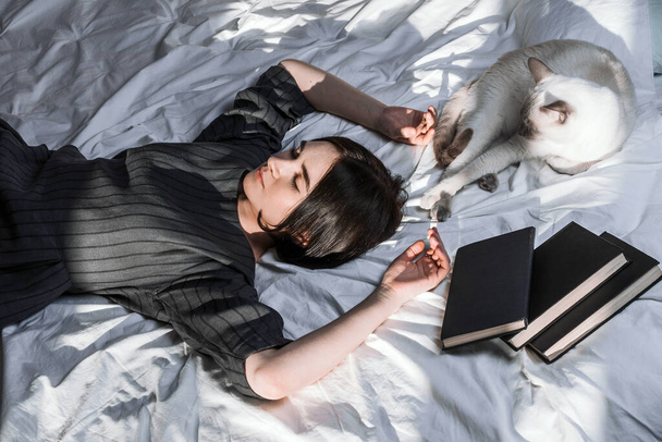 Young adult woman lying in bed and with her cat and pile of books in her hands, selective focus - Fotografie, Obrázek