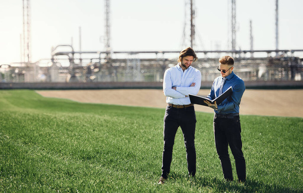 Two young engineers standing outdoors by oil refinery, discussing issues. - Fotoğraf, Görsel