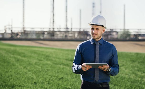 Young engineer with hard hat and tablet standing outdoors by oil refinery. - Foto, imagen
