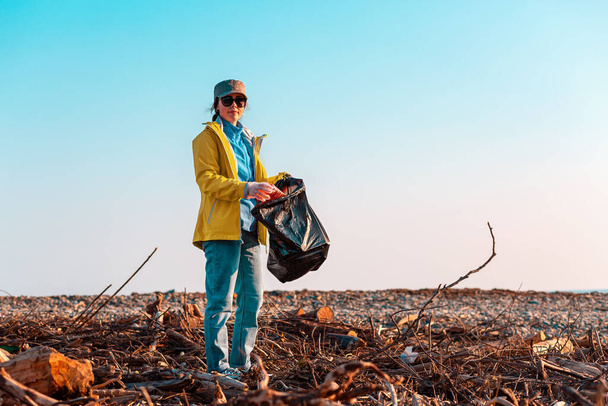 Une jeune bénévole caucasienne dans une veste verte met une bouteille en plastique dans un sac poubelle. En arrière-plan, une plage boueuse et le ciel. Concept de pollution de l'environnement. Espace de copie
. - Photo, image