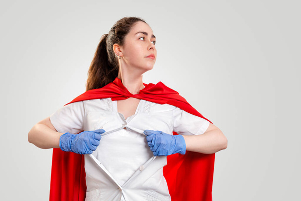 Een vrouwelijke arts, met medische handschoenen en een rode superheld Cape, scheurt haar jas op haar borst. Grijze achtergrond. Het concept van de Kracht van een superheld voor de geneeskunde. - Foto, afbeelding