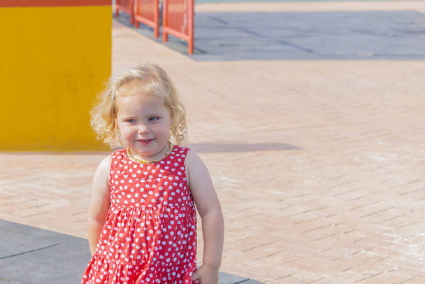 Nettes kleines Mädchen in rot gepunktetem Kleid, das auf dem Spielplatz spaziert. Ein kleines Mädchen läuft auf dem Spielplatz auf der Straße. Kopierraum. Sommerfreizeit und glückliche Kindheit - Foto, Bild