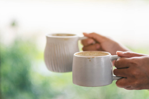 Imagen de primer plano de las manos sosteniendo dos tazas blancas de café caliente con fondo de naturaleza borrosa
 - Foto, imagen